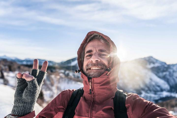 Mann mit Outdoor-Ausrüstung beim Bergwandern hebt gutgelaunt die Hand zum Gruß in die Kamera