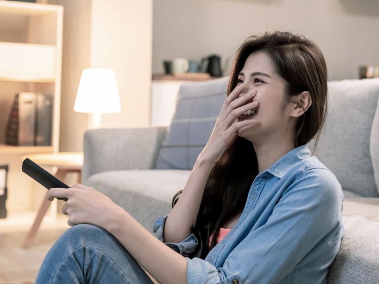 Eine Frau sitzt auf dem Boden ihres Wohnzimmers, hält eine Fernbedienung in der Hand und schaut lächelnd TV.