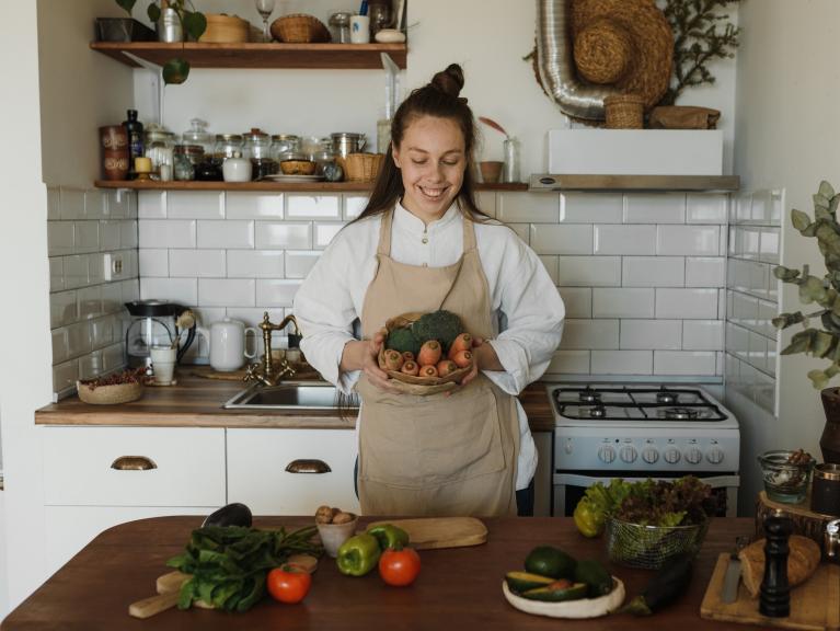 Eine Frau steht in einer Küche, lächelt und hält viel Gemüse in ihren Händen.