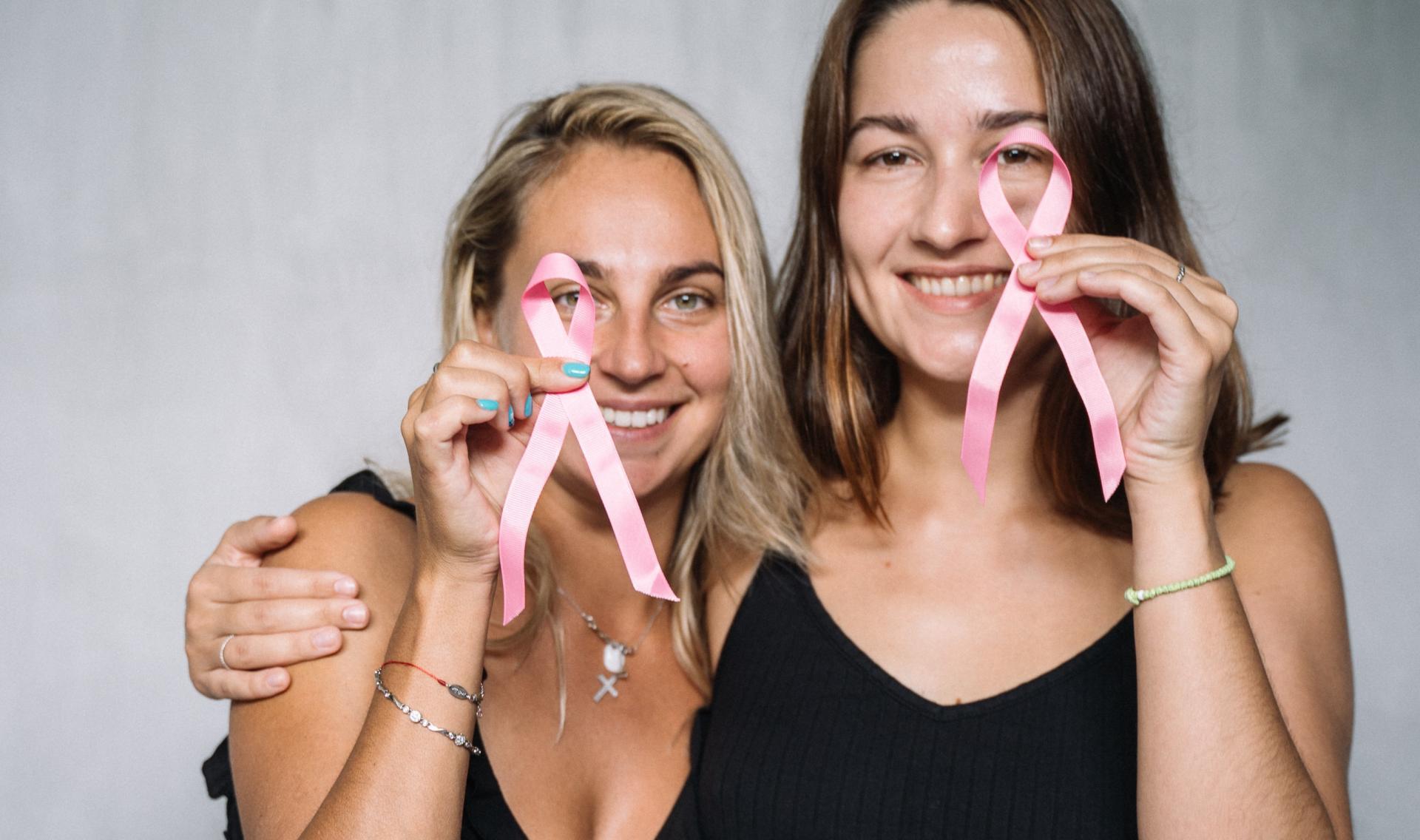 Zwei Frauen stehen Arm in Arm nebeneinander, lächeln und halten zwei rosa Krebs Ribbons. Rosa Krebs Ribbons stehen für Brustkrebs.