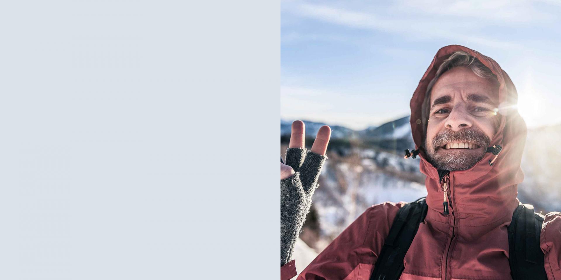 Mann mit Outdoor-Ausrüstung beim Bergwandern hebt gutgelaunt die Hand zum Gruß in die Kamera