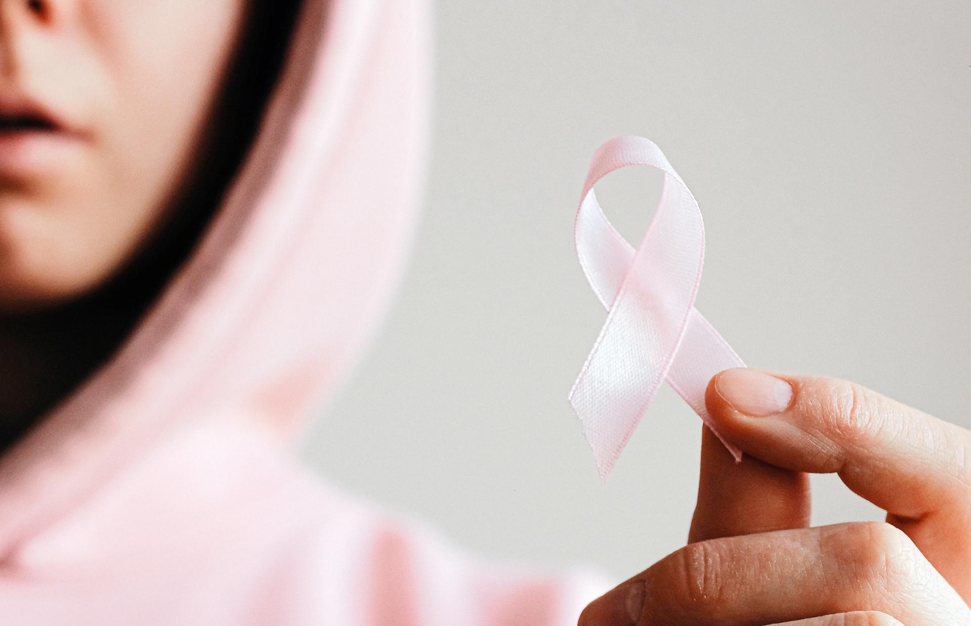 Eine Frau in einem rosafarbenen Pullover hält eine rosa Schleife in ihrer Hand, was das Zeichen für Brustkrebs darstellt.