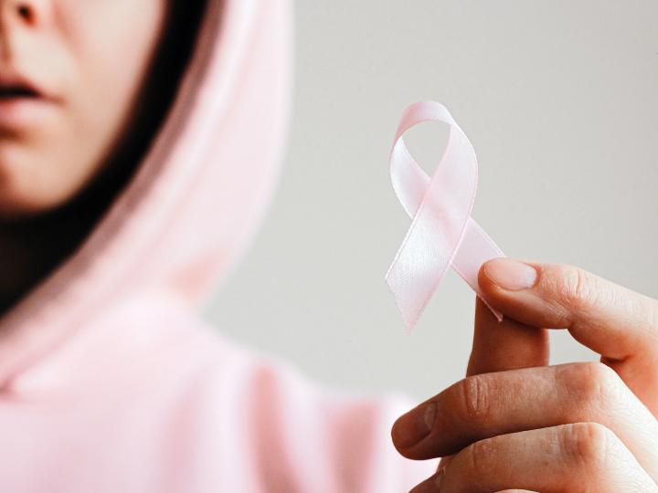 Eine Frau in einem rosafarbenen Pullover hält eine rosa Schleife in ihrer Hand, was das Zeichen für Brustkrebs darstellt.