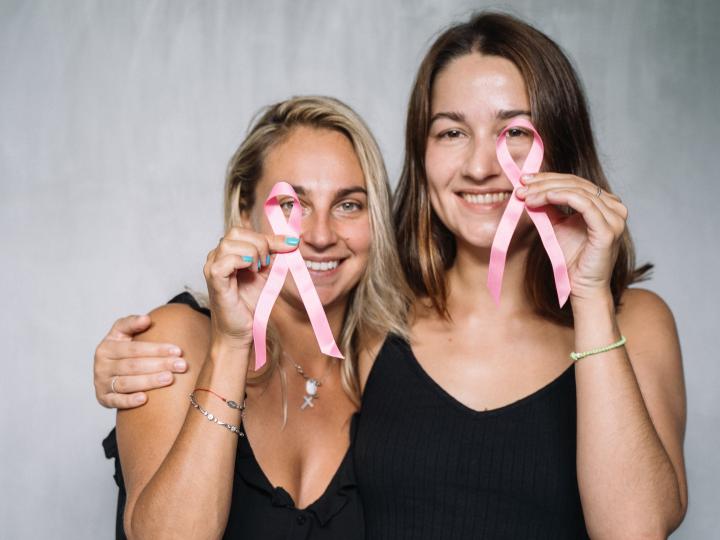 Zwei Frauen stehen Arm in Arm nebeneinander, lächeln und halten zwei rosa Krebs Ribbons. Rosa Krebs Ribbons stehen für Brustkrebs.